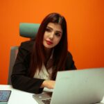 Focused businesswoman working on a laptop at a modern office desk with vibrant orange background.